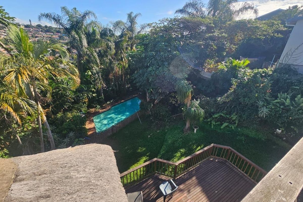 Elevated view of a lush garden featuring a swimming pool, a timber deck with balustrades, and a thatched roof section.