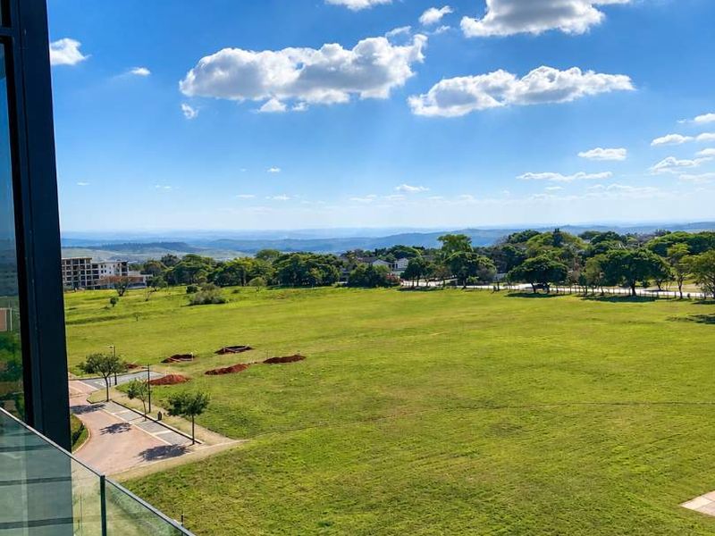 A clear, elevated view overlooking a large green field and surrounding landscape, captured from a balcony with a modern glass balustrade.