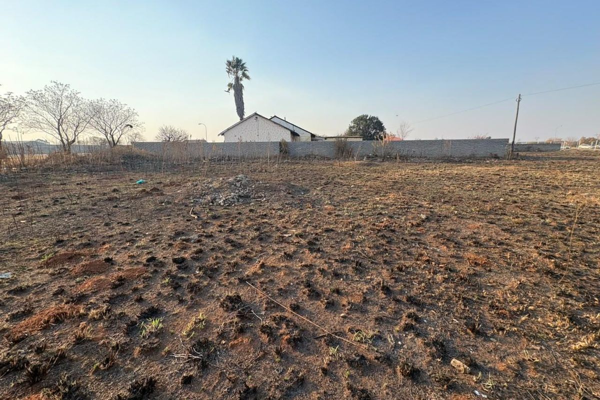 A view of a vacant residential plot featuring dry, uneven ground, a perimeter wall, and a utility pole in the background.