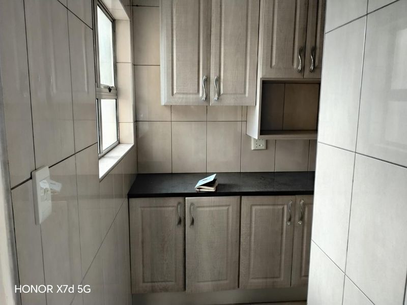 A compact scullery or utility area featuring light wood-finish cabinetry, dark countertops, and fully tiled walls with a functional window.