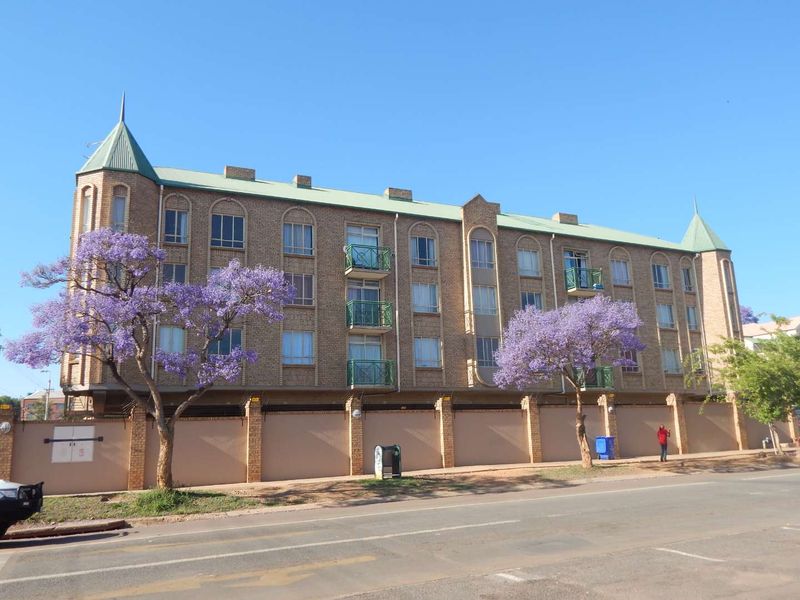 Exterior view of a multi-story brick apartment building featuring turret-style roof accents, balconies with metal railings, and a secure perimeter boundary wall.