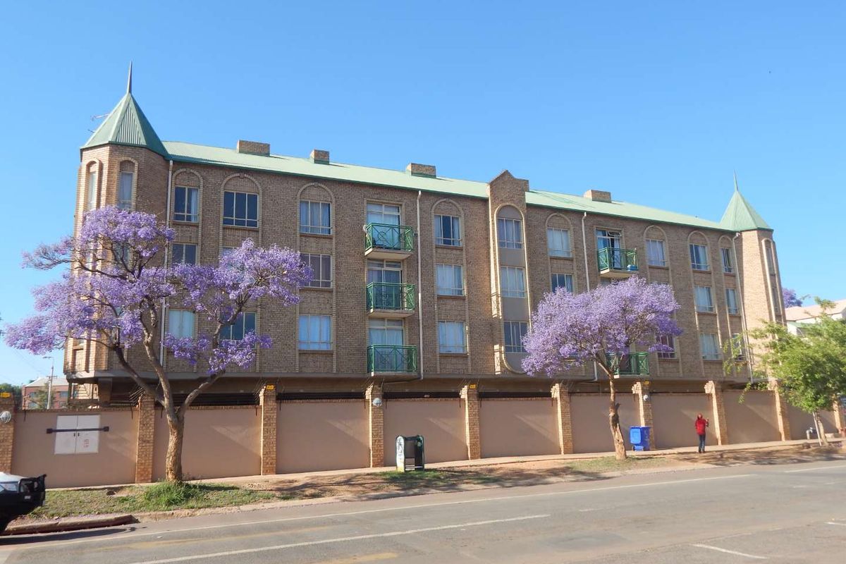 Exterior view of a multi-story brick apartment building featuring turret-style roof accents, balconies with metal railings, and a secure perimeter boundary wall.
