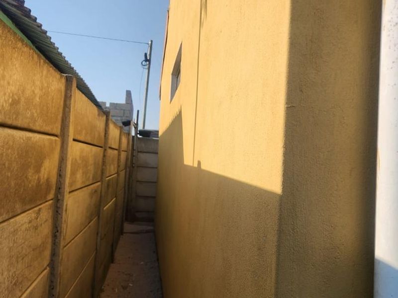 A narrow side passage between the main house structure and a precast concrete boundary wall, featuring a plastered exterior wall and a downpipe.
