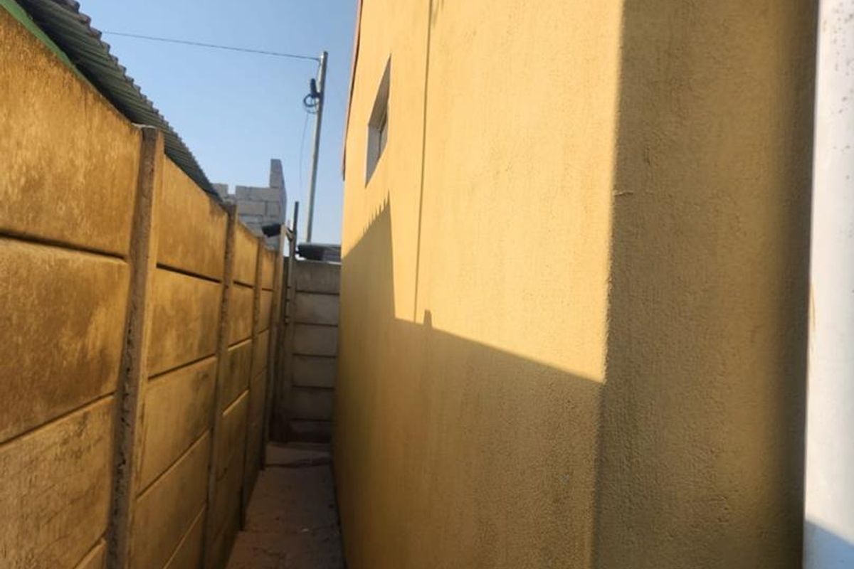 A narrow side passage between the main house structure and a precast concrete boundary wall, featuring a plastered exterior wall and a downpipe.