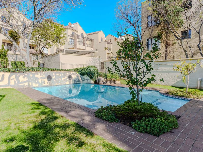 A well-maintained outdoor swimming pool area featuring brick paving, landscaped garden beds, and a retaining wall within a residential apartment complex.