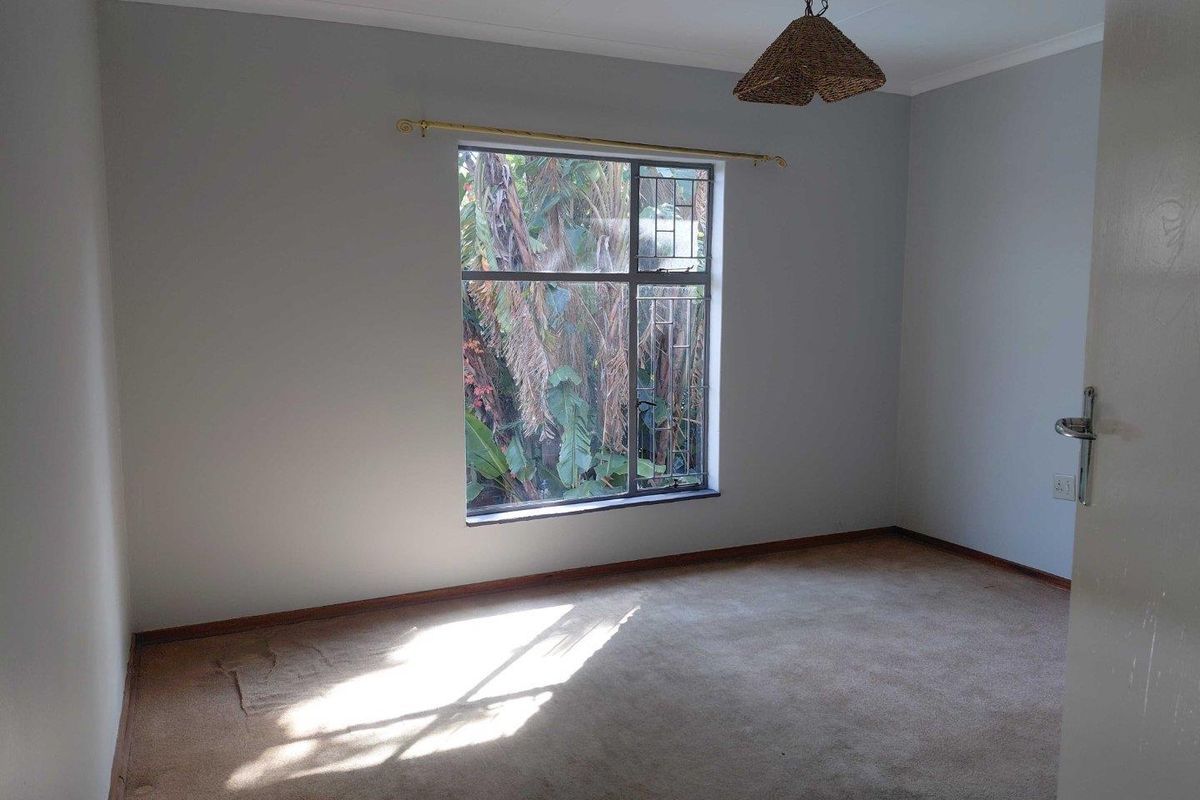 A vacant bedroom featuring neutral walls, carpeted flooring, and a large window with security bars overlooking garden greenery.