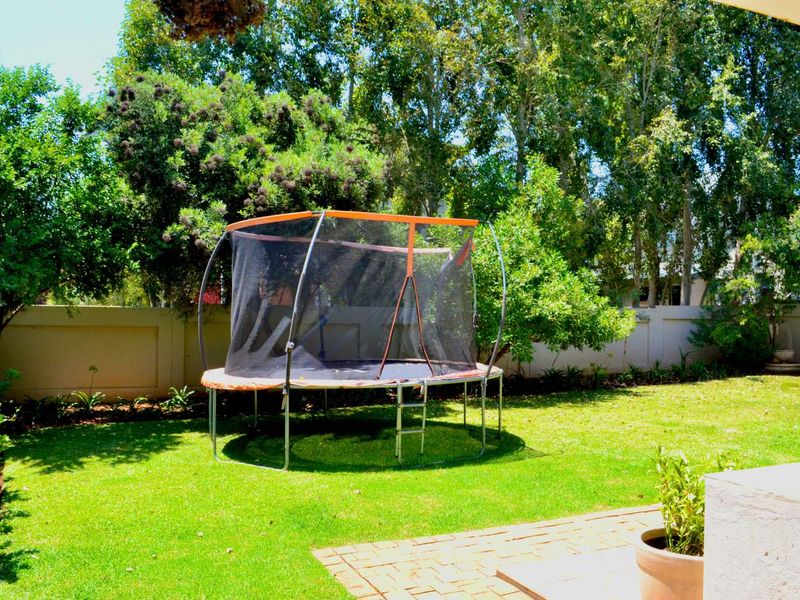 A well-maintained private garden featuring a lush green lawn, mature trees, and a perimeter boundary wall with an adjacent paved patio area.
