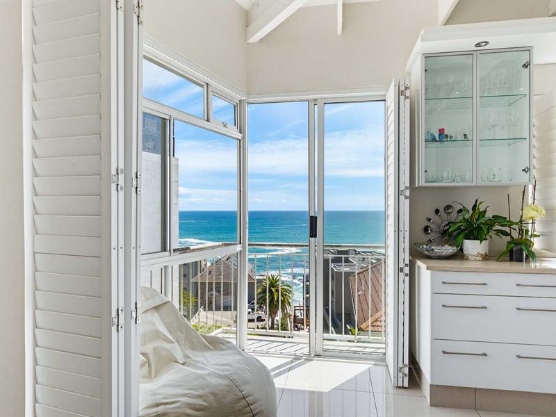 A bright, modern balcony area featuring white plantation shutters, built-in cabinetry with glass display shelving, and expansive ocean views through sliding glass doors.