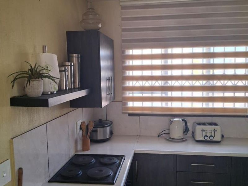 A functional kitchen corner featuring dark cabinetry, white countertops, a built-in electric hob, tiled backsplash, and a window with modern roller blinds.