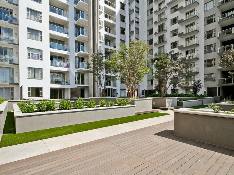 A well-maintained communal courtyard area featuring raised garden planters, composite decking, and paved walkways surrounded by modern apartment building facades with glass-fronted balconies.