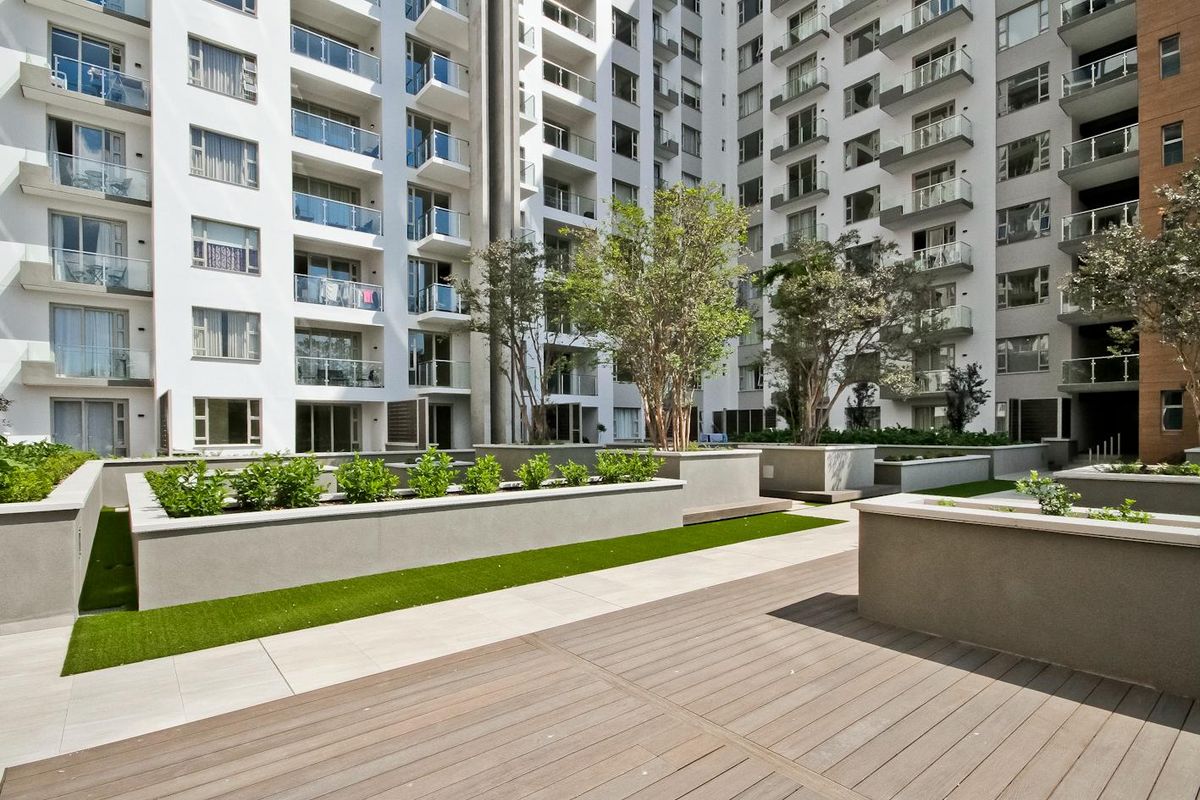 A well-maintained communal courtyard area featuring raised garden planters, composite decking, and paved walkways surrounded by modern apartment building facades with glass-fronted balconies.