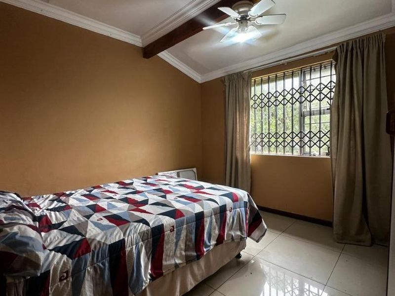 A bedroom featuring tiled flooring, a ceiling fan with an exposed wooden beam, and a window fitted with a security gate.