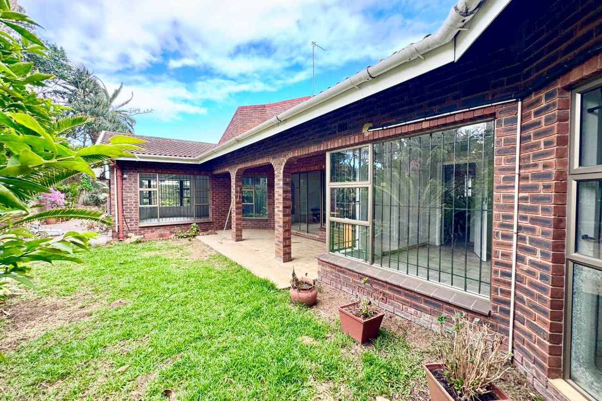 Rear view of the house featuring a brick facade, covered patio area, and large windows with security bars overlooking the garden.