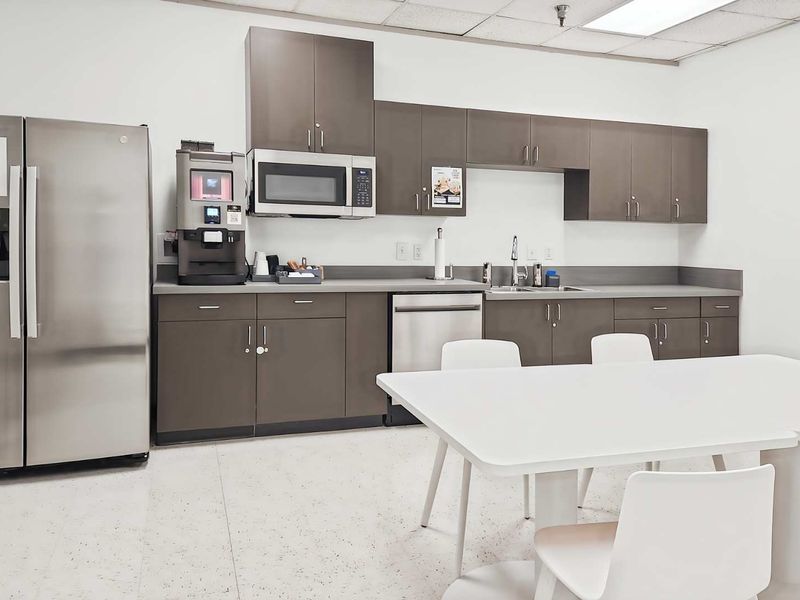 A clean office kitchenette featuring modern grey cabinetry, stainless steel appliances, a sink, and a drop ceiling with integrated lighting.