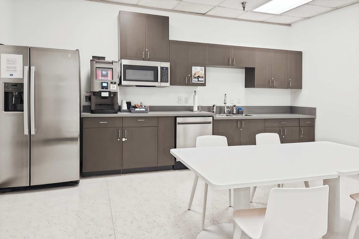 A clean office kitchenette featuring modern grey cabinetry, stainless steel appliances, a sink, and a drop ceiling with integrated lighting.