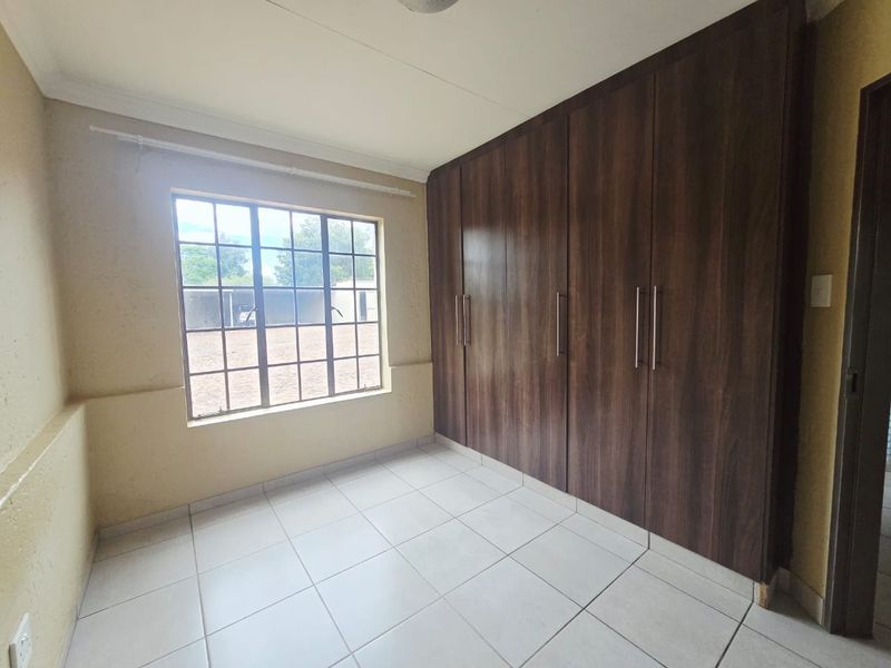 A clean, empty bedroom featuring neutral tiled flooring, large built-in dark wood cupboards, and a window with security bars.