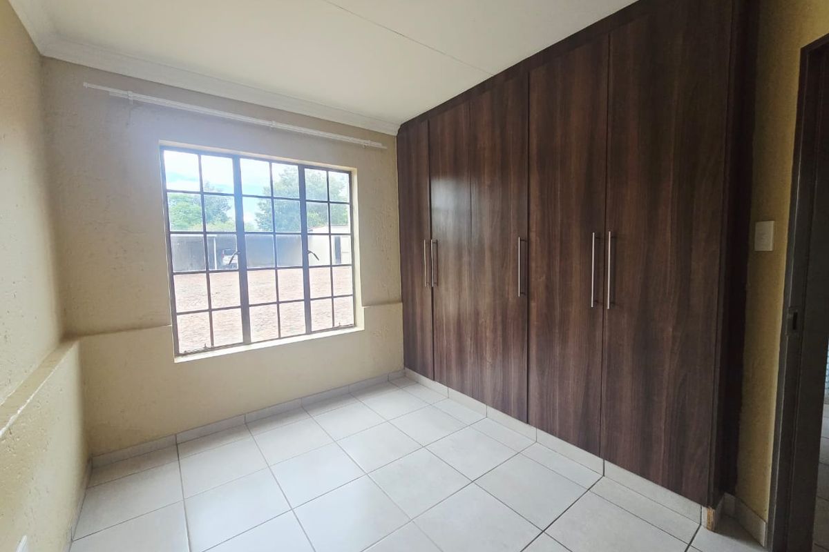 A clean, empty bedroom featuring neutral tiled flooring, large built-in dark wood cupboards, and a window with security bars.