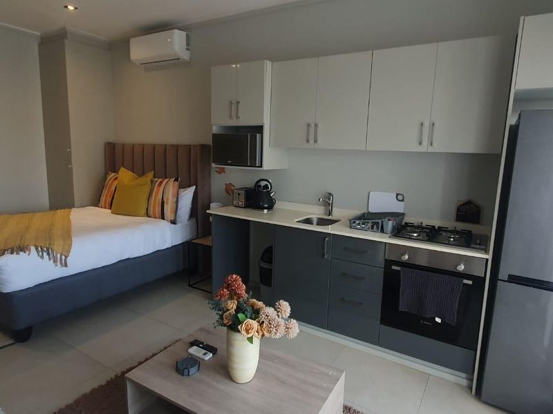 A modern open-plan studio apartment featuring a compact kitchenette with built-in appliances, tiled flooring, and an integrated air conditioning unit.