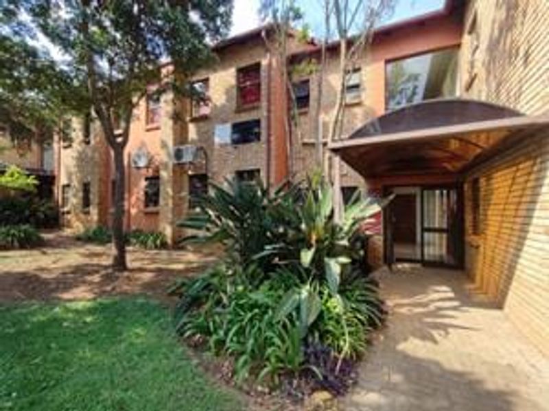 Exterior view of the apartment building entrance featuring a brick facade, covered entryway, and surrounding landscaped garden area.