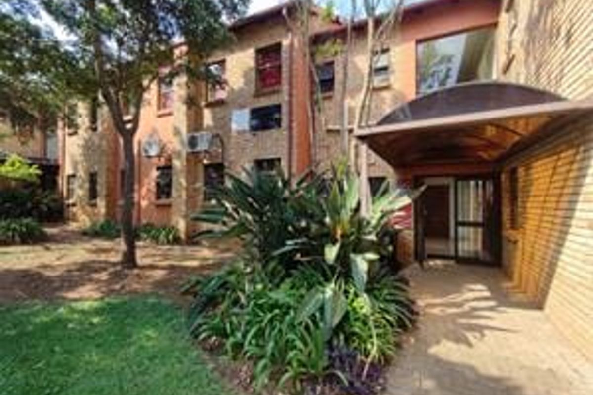 Exterior view of the apartment building entrance featuring a brick facade, covered entryway, and surrounding landscaped garden area.