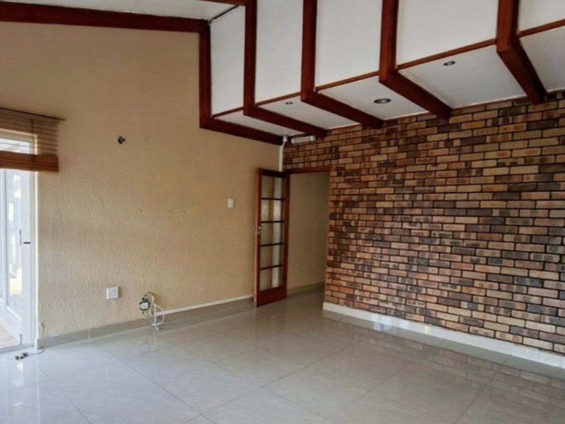 Spacious living area featuring an exposed brick feature wall, timber ceiling beams, and tiled flooring, offering a versatile layout for modern living.