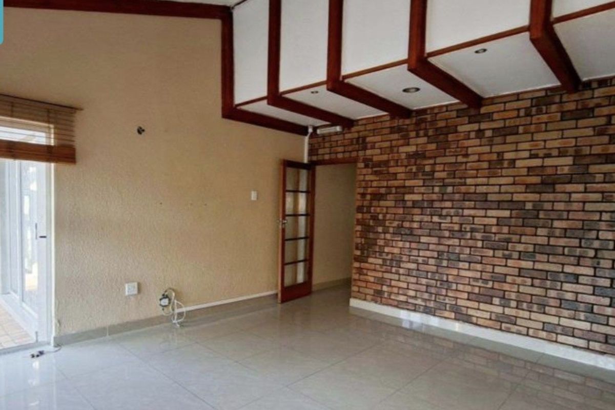 Spacious living area featuring an exposed brick feature wall, timber ceiling beams, and tiled flooring, offering a versatile layout for modern living.