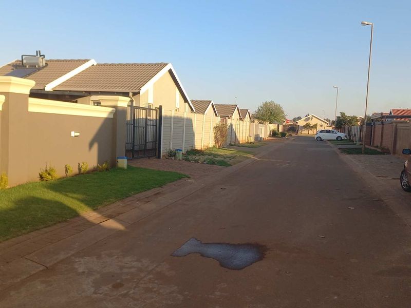 A street view showing the residential neighbourhood approach with perimeter walls, gated access, and tiled roof houses.