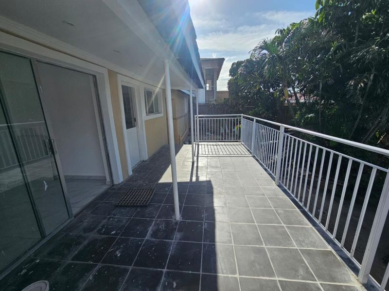 A spacious covered balcony featuring tiled flooring and a white metal balustrade, offering an open outdoor area accessible via sliding glass doors.