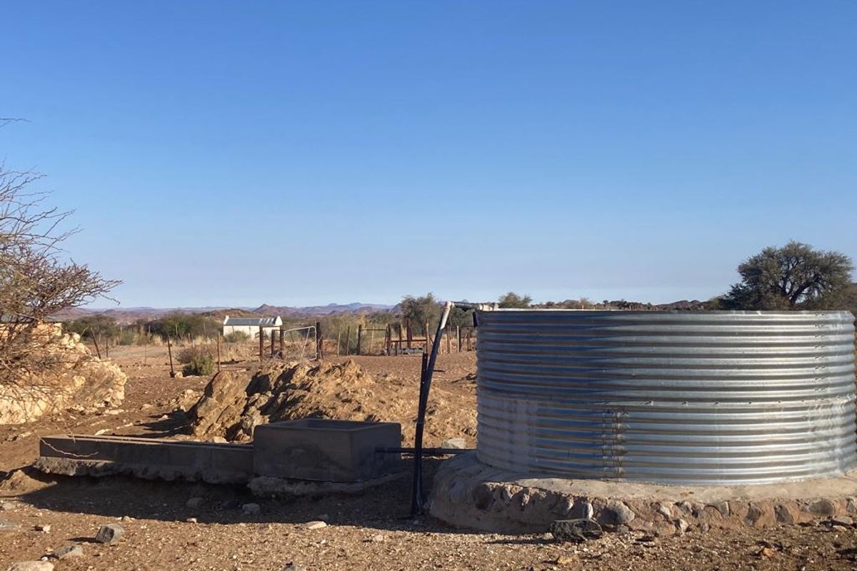 A corrugated metal water storage tank with an attached concrete livestock trough, situated in an open farm landscape with distant mountain views.