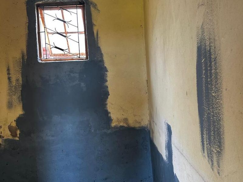 A small, vacant utility space featuring concrete flooring, a window with security bars, and walls requiring significant repair and repainting.