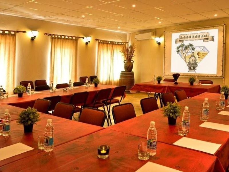 A well-lit hotel conference or event room featuring a projector screen, wall-mounted lighting, and air conditioning, set up with multiple tables for meetings.