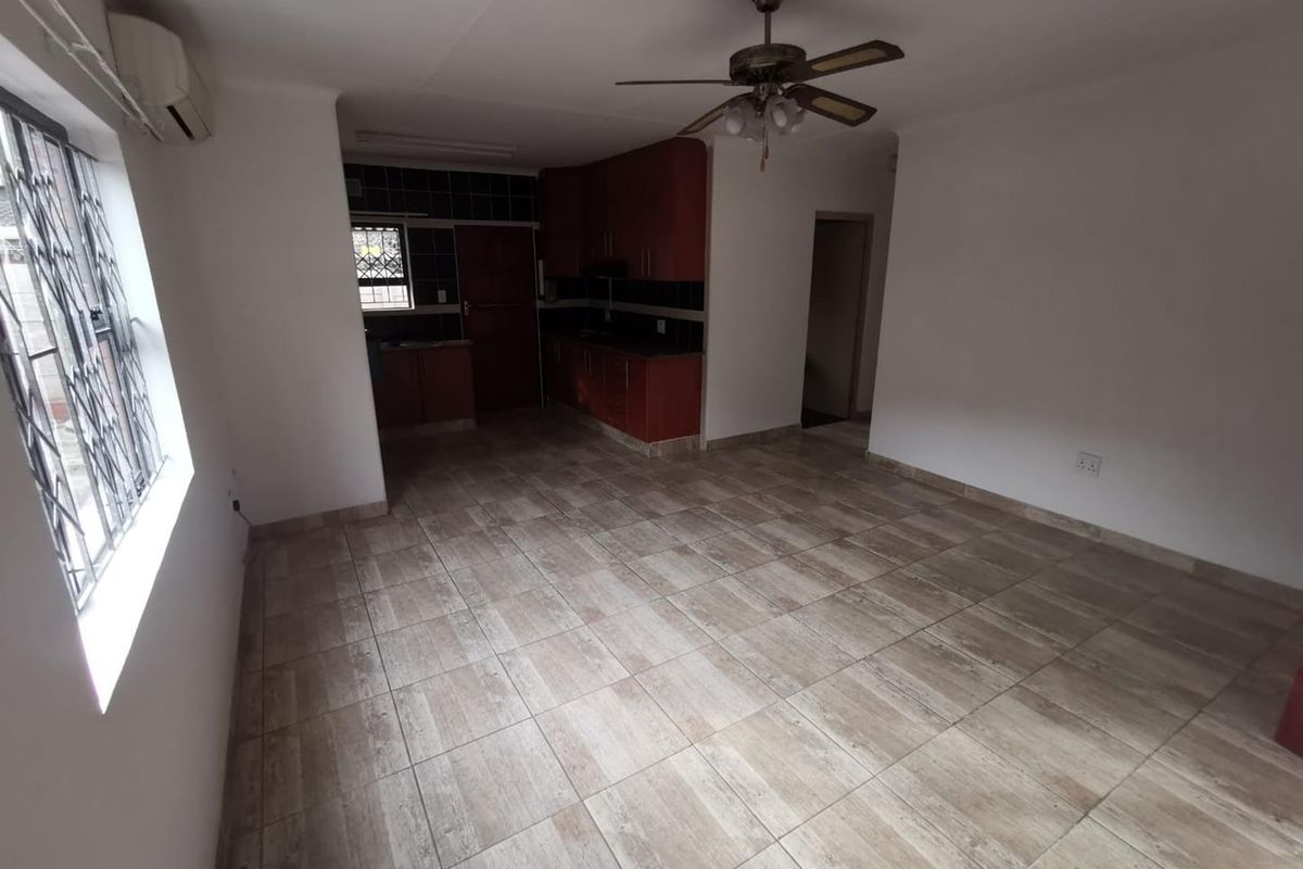 An open-plan living and kitchen area featuring tiled flooring, a ceiling fan, air conditioning, and built-in kitchen cabinetry with security bars on the windows.