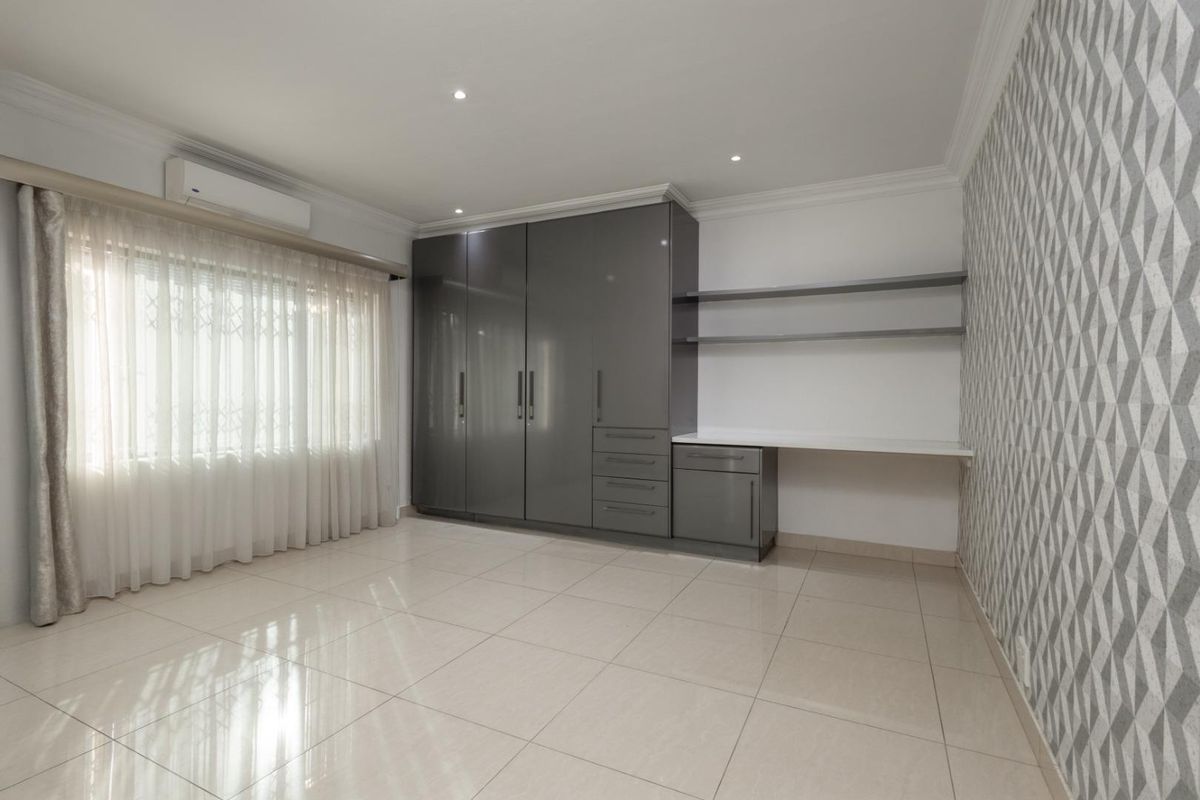 A bright, modern bedroom featuring high-gloss built-in cupboards, a matching integrated desk, tiled flooring, and a stylish geometric wallpaper feature wall.