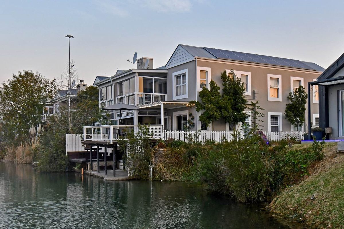 Rear view of a townhouse featuring a raised wooden deck, covered patio, white picket fencing, and direct waterfront access with solar panels on the roof.