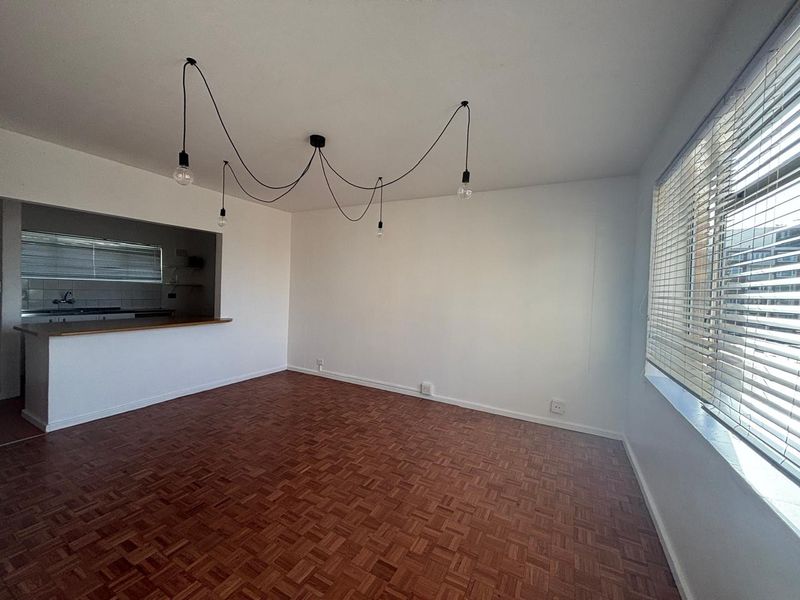 An open-plan living area featuring classic parquet flooring, a breakfast bar overlooking the kitchen, and large windows with fitted venetian blinds.