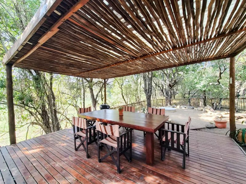 A covered outdoor patio area featuring timber decking and a rustic thatched pergola roof, set within a natural wooded garden environment.