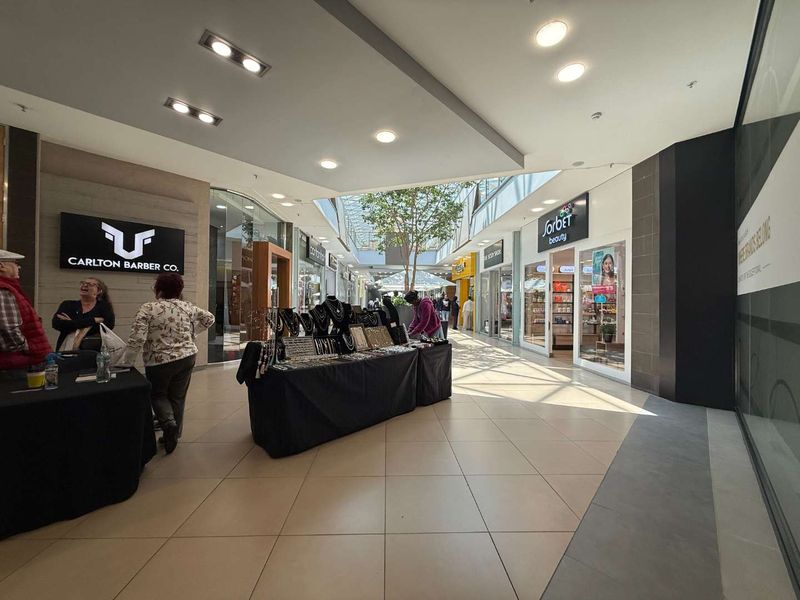 A bright, modern retail shopping centre corridor featuring tiled floors, recessed lighting, a central skylight, and multiple glass-fronted retail storefronts.