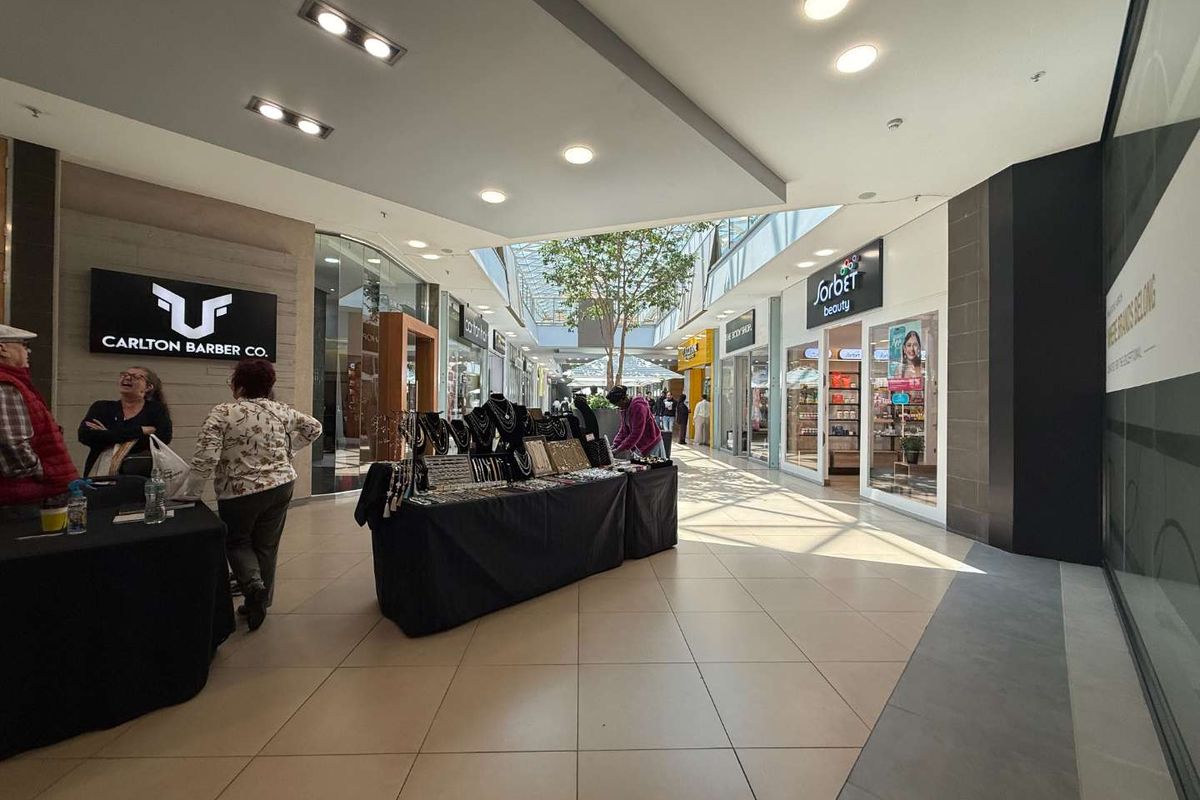 A bright, modern retail shopping centre corridor featuring tiled floors, recessed lighting, a central skylight, and multiple glass-fronted retail storefronts.