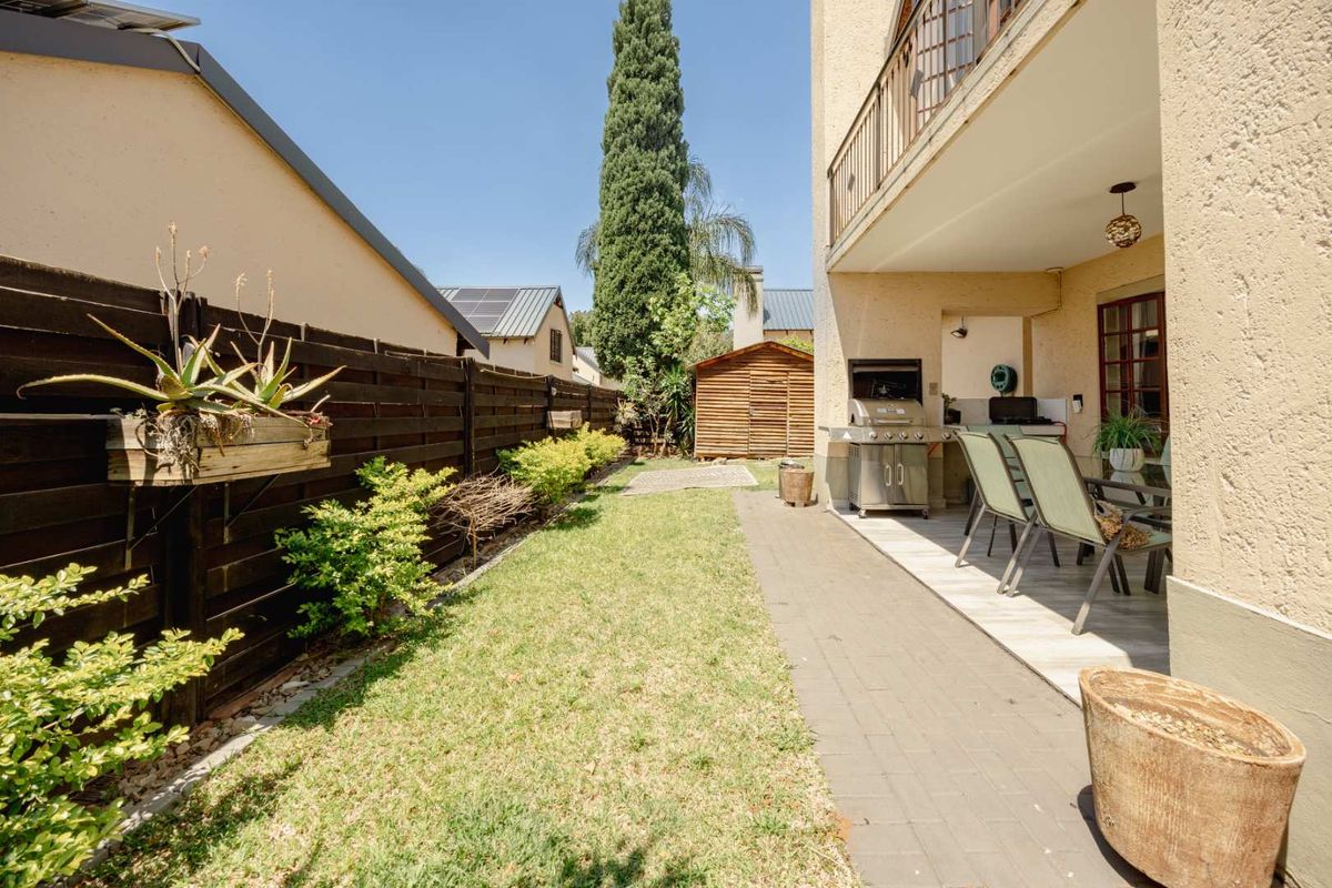 Rear exterior view featuring a covered patio with a built-in braai, paved walkway, lawn, and timber fencing.