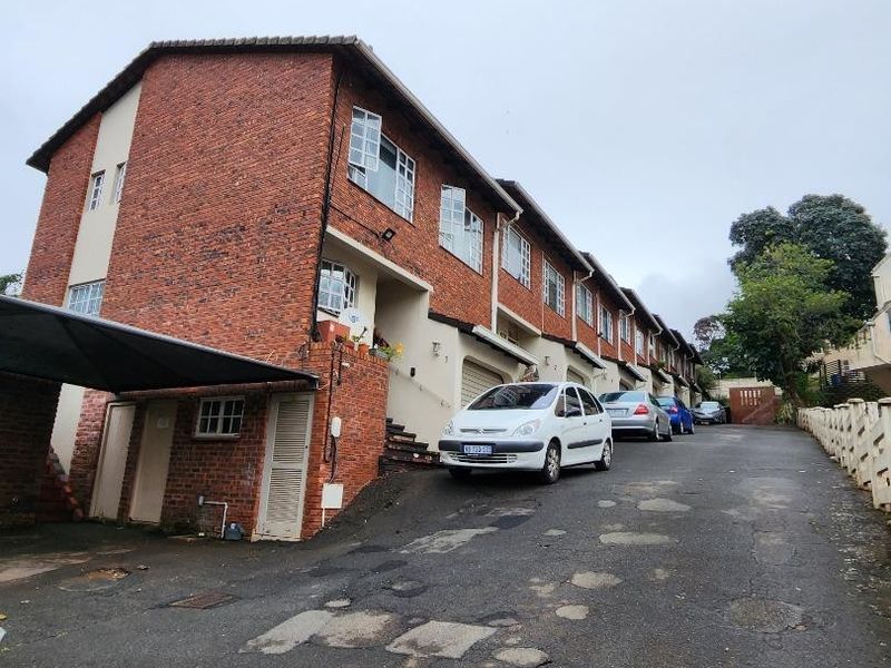 Exterior view of a multi-story face brick apartment complex featuring a shared asphalt driveway and covered parking areas.