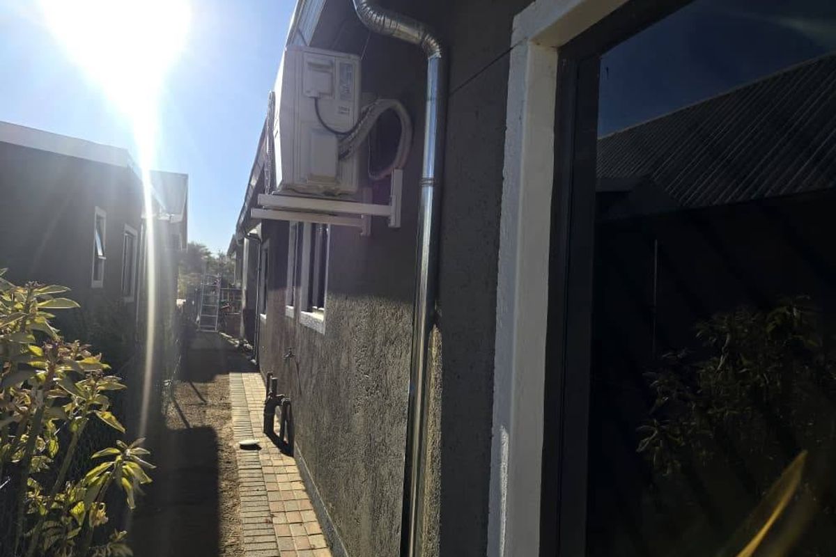 Side exterior view of the townhouse featuring a paved walkway, external air conditioning unit, and downpipe along the dark-rendered wall.