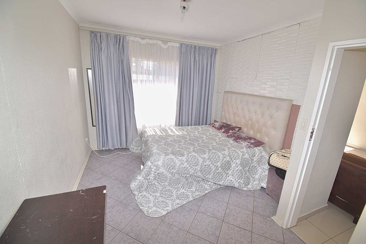 A bedroom featuring tiled flooring, a textured feature wall, and large windows with curtains, providing a functional and well-lit sleeping space.