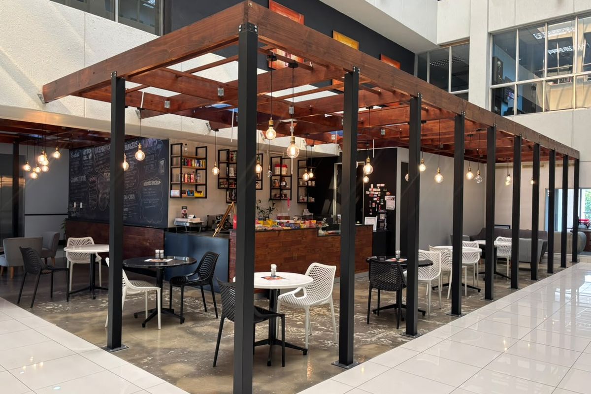 Modern open-plan office cafe area featuring a timber pergola structure, polished concrete floors, and integrated service counter with pendant lighting.