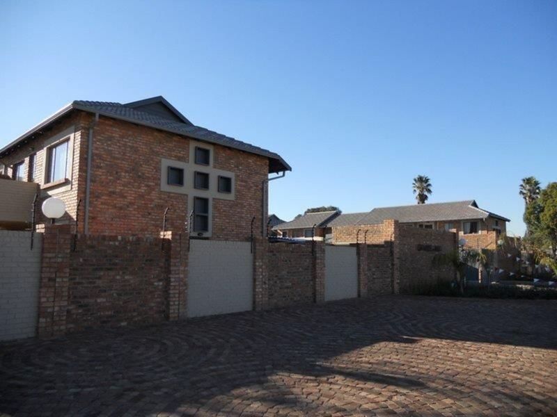 Front exterior view of a face brick townhouse complex featuring a paved driveway, perimeter wall with electric fencing, and distinctive architectural window detailing.