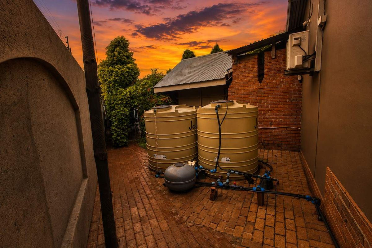 Side yard area featuring two large water storage tanks, a pool filtration pump, and brick paving, set against a sunset backdrop.