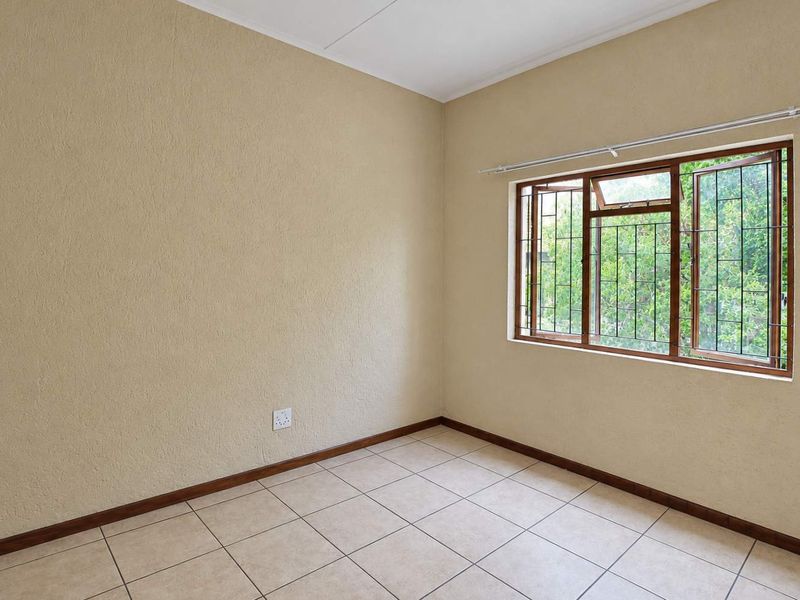 A bright, empty bedroom featuring neutral textured walls, tiled flooring, and a window fitted with security burglar bars.
