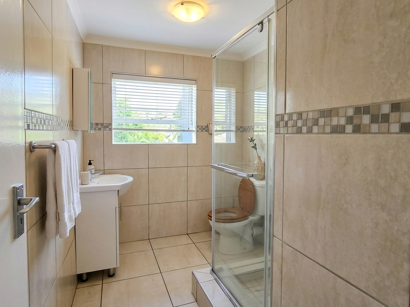 A clean, well-maintained bathroom featuring a glass shower enclosure, vanity basin, toilet, and neutral floor-to-ceiling wall tiles.