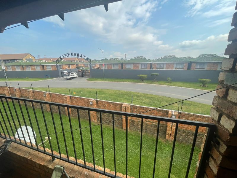 A view from an elevated balcony overlooking a residential complex entrance, featuring a metal balustrade and brick boundary wall with electric fencing.