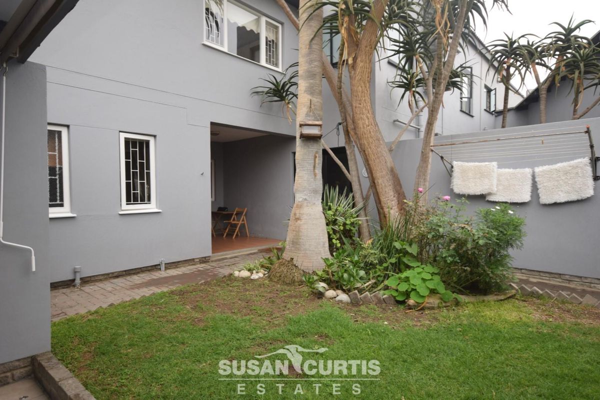 A rear exterior view of the property featuring a small lawn, established trees, and a paved walkway leading to the building entrance.