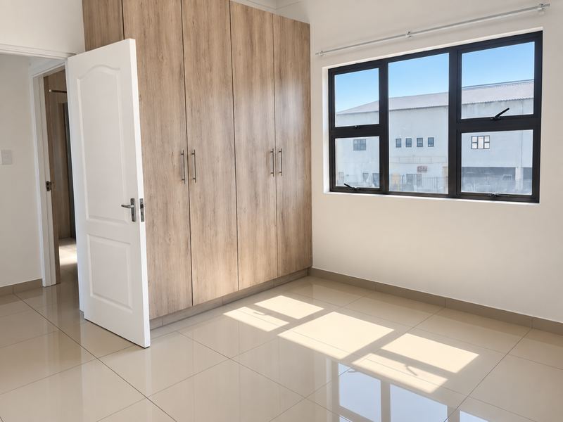 A bright, empty bedroom featuring modern wood-finish built-in wardrobes, neutral tiled flooring, and large aluminium-framed windows allowing for ample natural light.
